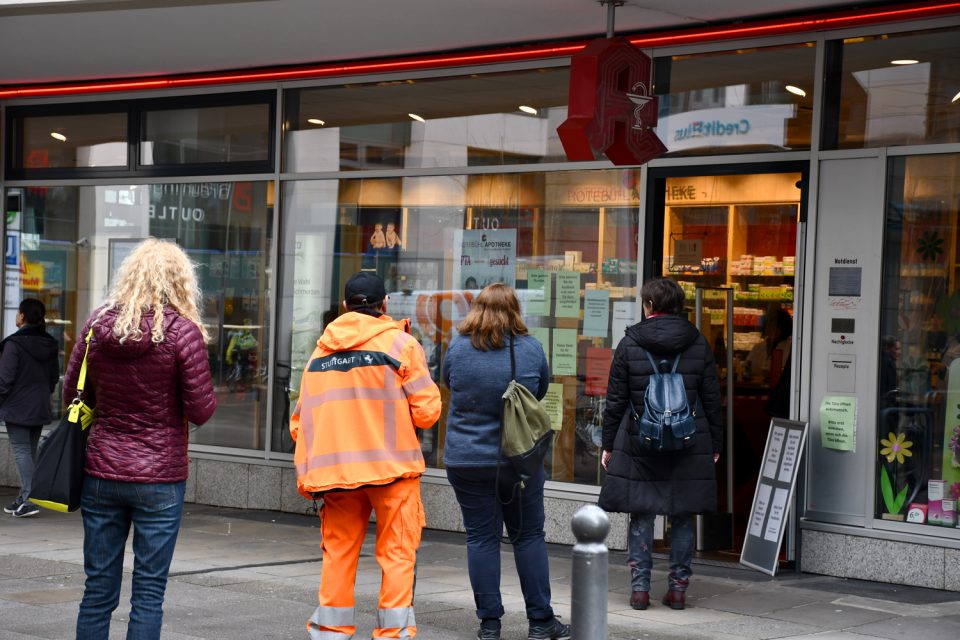 Vier Leute stehen in großem Abstand zueinander vor einer Apotheke in der Schlange. Einer der Wartenden trägt eine orangene Kluft, wie sie die Müllabfuhr trägt. Auf seinem Rücken ist das Logo der Stadt Stuttgart zu sehen.